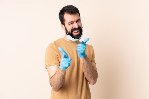 Hombre caucásico con barba protegiendo con una máscara y guantes sobre pared aislada apuntando hacia el frente y sonriendo