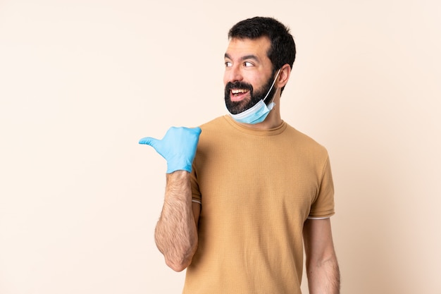 Hombre caucásico con barba protegiendo con una máscara y guantes sobre pared aislada apuntando hacia un lado para presentar un producto