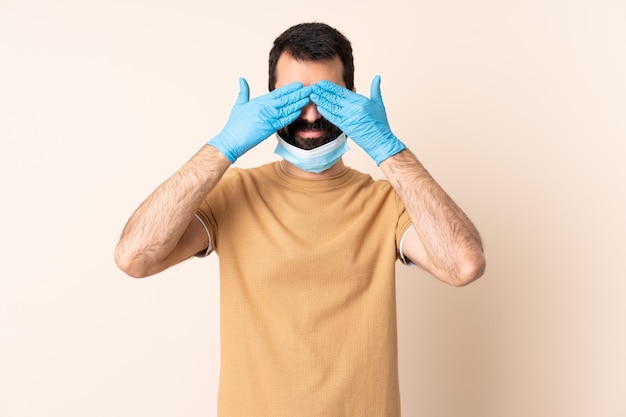 Hombre caucásico con barba protegiendo con una máscara y guantes sobre la pared aislada que cubre los ojos con las manos y sonriendo