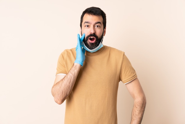 Hombre caucásico con barba protegiendo con una máscara y guantes sobre la pared con expresión facial sorprendida y conmocionada