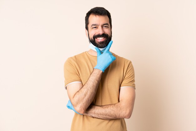 Hombre caucásico con barba protegiendo con una máscara y guantes sobre pared feliz y sonriente