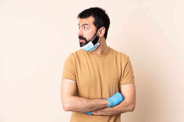 Hombre caucásico con barba protegiendo con una máscara y guantes sobre la pared mirando hacia un lado