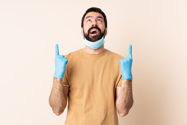 Hombre caucásico con barba protegiendo con una máscara y guantes sobre la pared sorprendido y apuntando hacia arriba