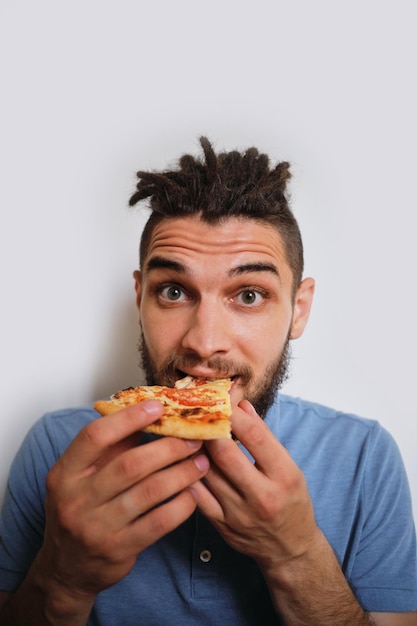 Hombre caucásico con barba y rastas come pizza sobre fondo blanco neutro Imagen vertical