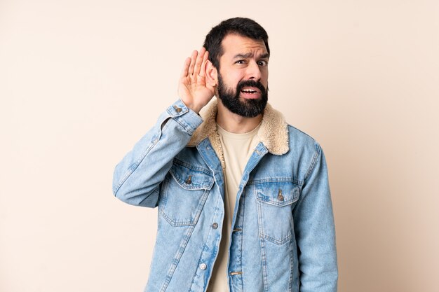 Hombre caucásico con barba sobre la pared escuchando algo poniendo la mano en la oreja