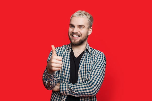 Hombre caucásico barbudo con cabello rubio está gesticulando el signo similar en una pared roja