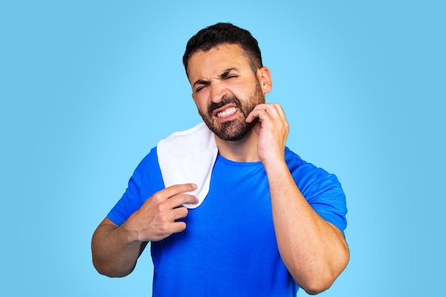 Un hombre caucásico barbudo con una camiseta azul con una toalla se rasca la barba con un fondo azul. ir