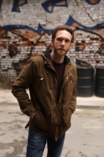 Hombre caucásico barbudo con una chaqueta marrón posando frente a un graffiti