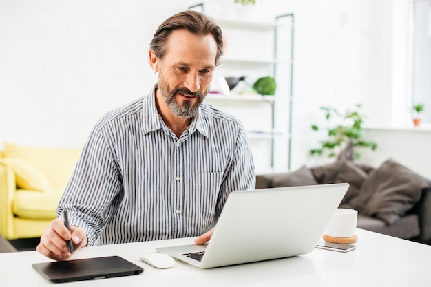 Hombre caucásico barbudo positivo sentado frente a su computadora portátil y sonriendo mientras sostiene el lápiz en la tableta gráfica