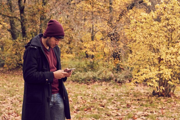 Hombre caucásico blanco en el bosque vestido con un abrigo largo y una gorra mirando el teléfono inteligente Colores de otoño