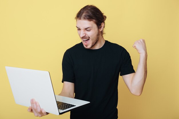 Hombre caucásico con cabello largo y barba está gesticulando el signo de sí posando en una pared amarilla sosteniendo una computadora portátil