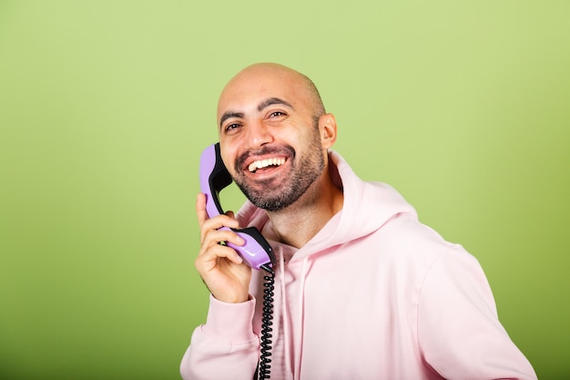 Hombre caucásico calvo joven en sudadera con capucha rosa aislado, sostenga el teléfono fijo con cara de asombro emocionado feliz