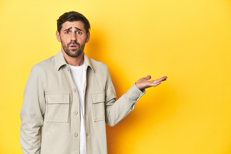 Foto hombre caucásico en camisa gris estudio disparado en amarillo impresionado sosteniendo espacio de copia en la palma