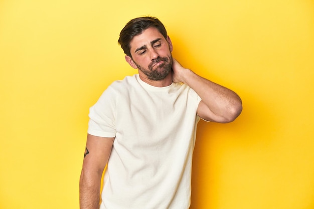 Hombre caucásico con camiseta blanca en fondo de estudio amarillo con dolor de cuello debido al estrés