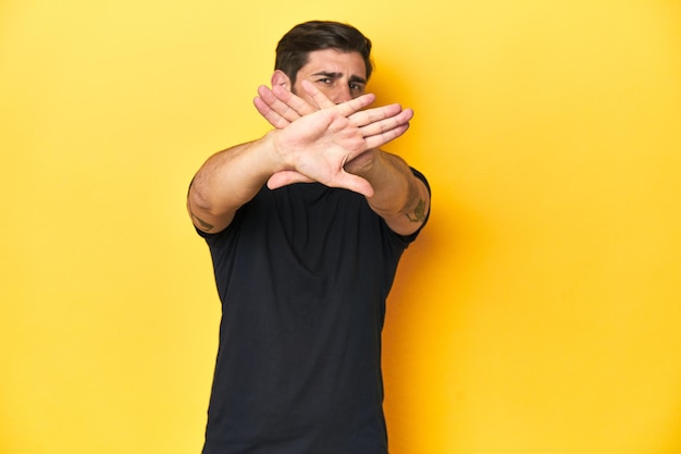 Foto hombre caucásico con camiseta negra y telón de fondo de estudio amarillo haciendo un gesto de negación