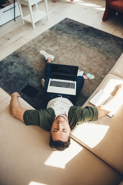 Hombre caucásico cansado con cerdas acostado en el piso y el sofá después de trabajar en la computadora portátil con un libro