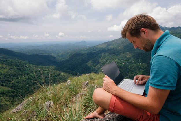 Hombre caucásico con una computadora portátil sentado en el borde de una montaña con impresionantes vistas trabajando en una computadora portátil Freelancer in Sri Lnaka