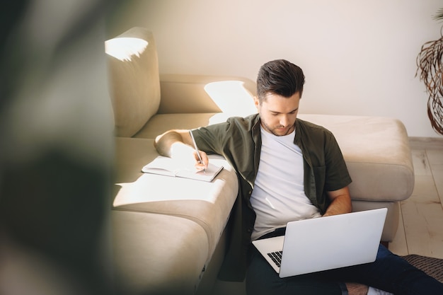 El hombre caucásico se concentra en trabajar en la computadora portátil desde su casa en el piso y escribir algo en el cuaderno