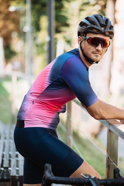 Hombre caucásico deportivo con casco de ropa deportiva y gafas posando al aire libre cerca de su bicicleta deportiva negra Un hombre guapo tomando un descanso después de andar en bicicleta al aire libre