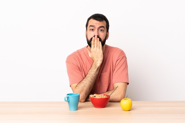 Hombre caucásico desayunando en una mesa cubriendo la boca con la mano.