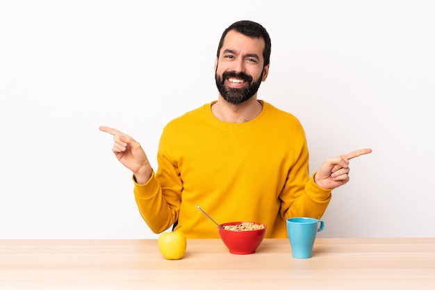 Hombre caucásico desayunando en una mesa señalando con el dedo a los laterales y feliz