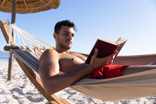 Hombre caucásico disfrutando del tiempo en la playa, tumbado en una hamaca y leyendo un libro