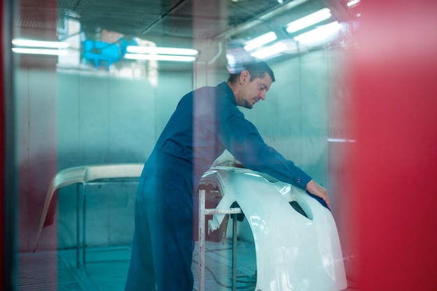 El hombre caucásico está limpiando piezas de automóviles antes de rociar color en el taller de pintura mecánica de reparación