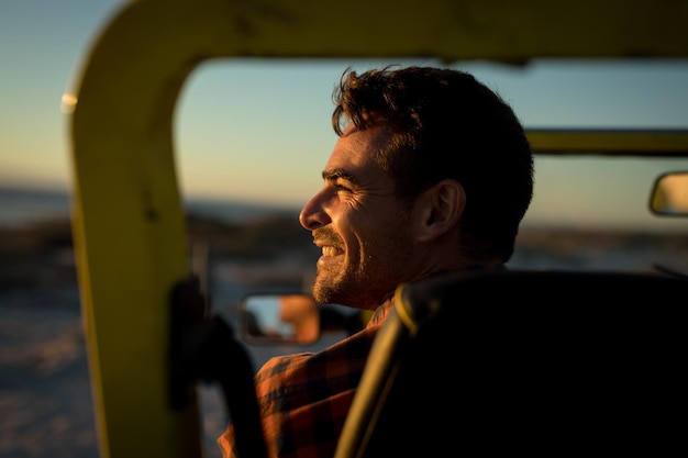 Hombre caucásico feliz sentado en un buggy de playa junto al mar durante la puesta de sol sonriendo. descanso en la playa en viaje de vacaciones de verano