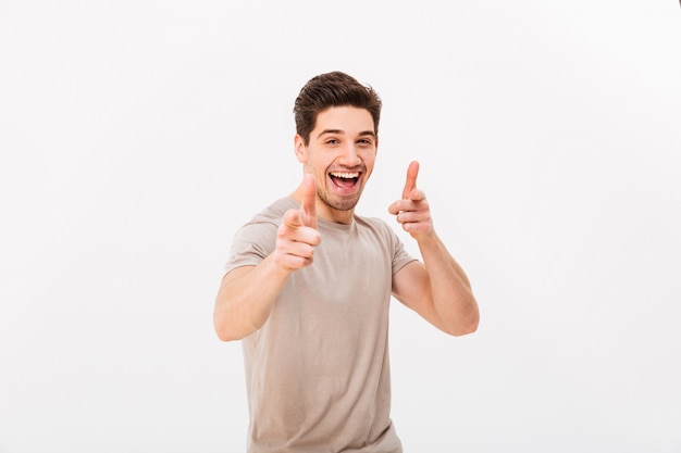 Hombre caucásico fresco en camiseta beige gesticulando con los dedos índices en la cámara, lo que significa hola o buena suerte, aislado sobre la pared blanca