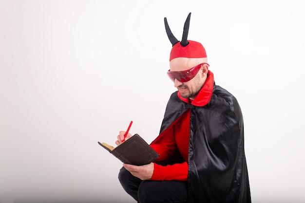 Hombre caucásico en gafas de sol rojas y disfraz de halloween con lápiz y cuaderno sobre fondo blanco de estudio.