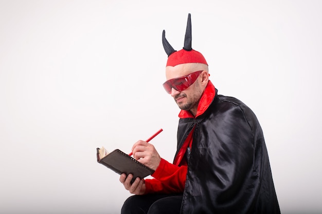 Hombre caucásico en gafas de sol rojas y disfraz de halloween con lápiz y cuaderno sobre fondo blanco de estudio.