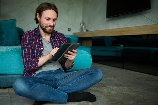 Hombre caucásico guapo y confiado con barba en jeans casuales y camisa a cuadros sosteniendo una tableta digital y mirando la cámara sentado en el suelo con la espalda contra el sofá mientras trabaja desde casa
