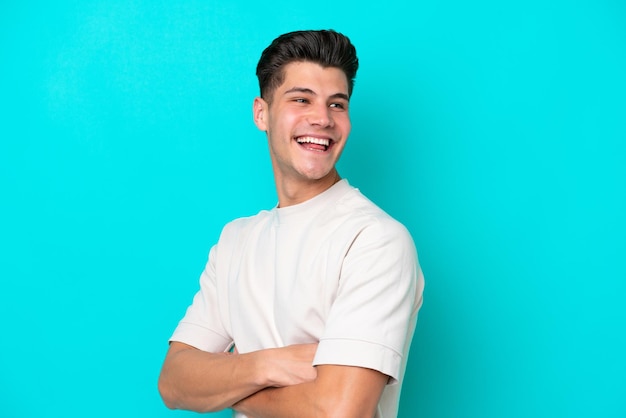 Foto hombre caucásico guapo joven aislado en bakcground azul con los brazos cruzados y feliz