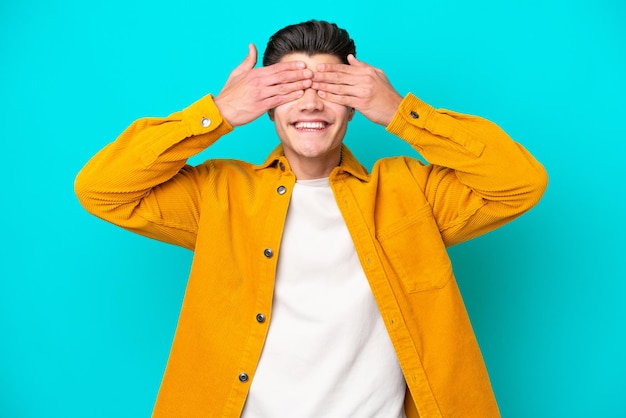 Hombre caucásico guapo joven aislado en bakcground azul cubriendo los ojos con las manos y sonriendo