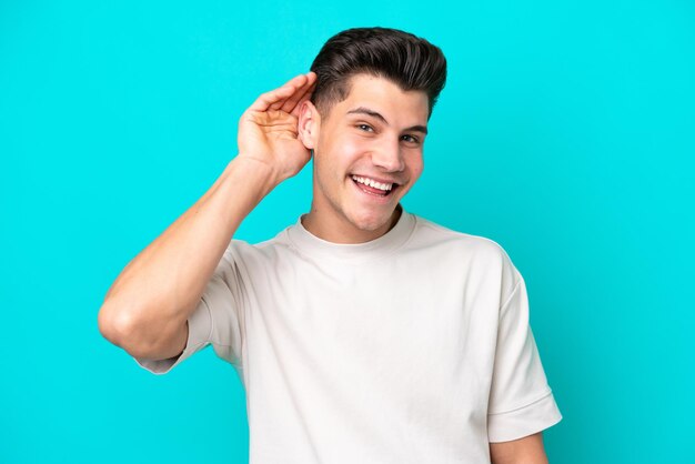 Hombre caucásico guapo joven aislado en bakcground azul escuchando algo poniendo la mano en la oreja