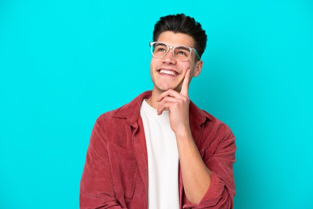 Hombre caucásico guapo joven aislado en bakcground azul con gafas y pensando mientras mira hacia arriba