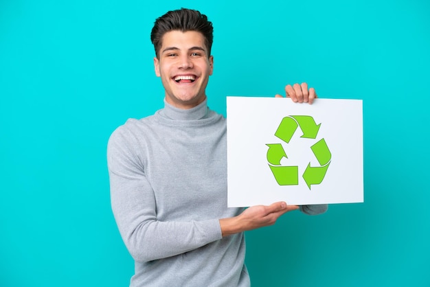 Hombre caucásico guapo joven aislado en bakcground azul sosteniendo un cartel con icono de reciclaje con expresión feliz