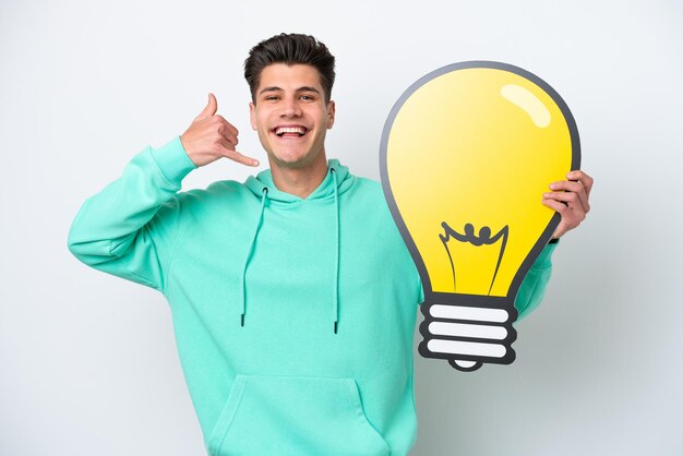 Hombre caucásico guapo joven aislado en bakcground blanco sosteniendo un icono de bombilla y haciendo gesto de teléfono