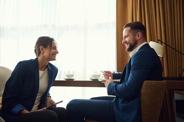 Hombre caucásico guapo y mujer atractiva, socios comerciales, asesores de inversiones vestidos con trajes de negocios sentados en un sillón y discutiendo asuntos comerciales en una habitación de hotel durante un viaje de negocios