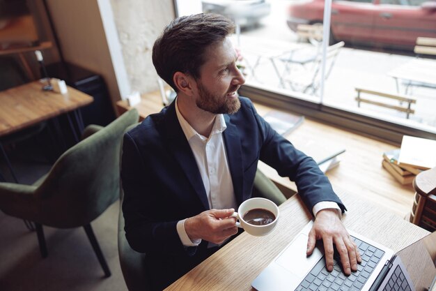 Hombre caucásico guapo sosteniendo una taza de café y sentado en la computadora portátil en una cafetería acogedora en la ventana
