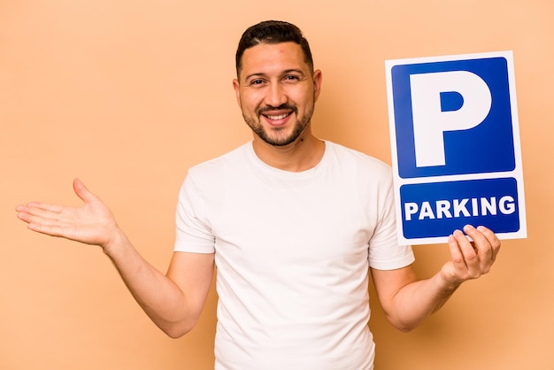 Hombre caucásico hispano sosteniendo un cartel de estacionamiento aislado en un fondo beige que muestra un espacio de copia en una palma y sosteniendo otra mano en la cintura