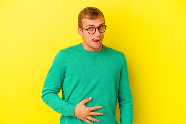 Hombre caucásico joven aislado en amarillo toca la barriga, sonríe suavemente, comiendo y concepto de satisfacción.