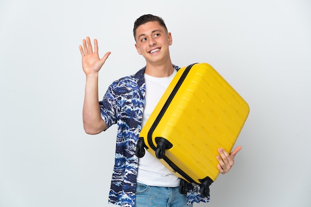 Hombre caucásico joven aislado en blanco en vacaciones con maleta de viaje y saludando