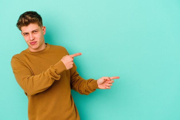 Hombre caucásico joven aislado en la pared azul apuntando con los dedos índices a un espacio de copia, expresando entusiasmo y deseo