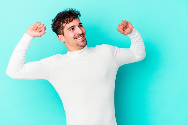 Hombre caucásico joven aislado en la pared azul celebrando un día especial, salta y levanta los brazos con energía.