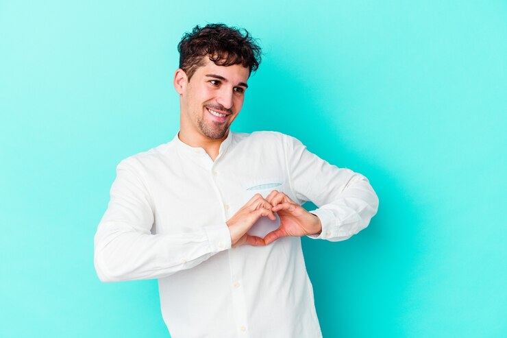 Foto hombre caucásico joven aislado en la pared azul sonriendo y mostrando una forma de corazón con las manos.
