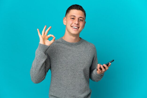 Hombre caucásico joven aislado en la pared azul con teléfono móvil y haciendo el signo de OK