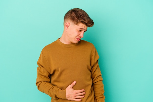 Hombre caucásico joven aislado en la pared azul toca la barriga, sonríe suavemente, comiendo y concepto de satisfacción.