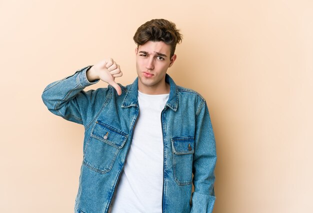 Hombre caucásico joven aislado en la pared beige mostrando el pulgar hacia abajo, concepto de decepción.