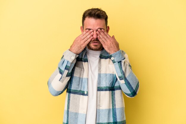 Hombre caucásico joven aislado sobre fondo amarillo miedo cubriendo los ojos con las manos